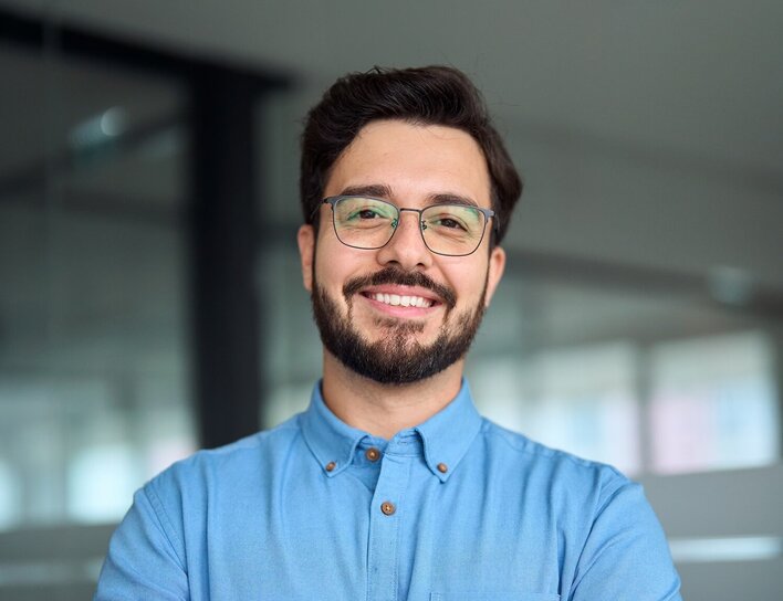 Titelbild Bachelor Betriebswirtschaft & Wirtschaftspsychologie Ein junger Mann mit Brille und Bart steht mit verschränkten Armen in einem modernen Büro und lächelt freundlich in die Kamera.