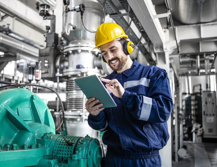 Titelbild Bachelor Verfahrenstechnik Ein Techniker mit Schutzhelm und Tablet inspiziert lächelnd Maschinen in einer modernen Industrieanlage.