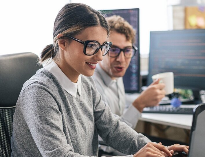Titelbild Bachelor IT-Management Eine junge Frau mit Brille arbeitet konzentriert an einem Laptop in einem modernen Büro. Ein Kollege mit Kaffeetasse sitzt daneben, während mehrere Bildschirme mit Code und Daten im Hintergrund zu sehen sind.