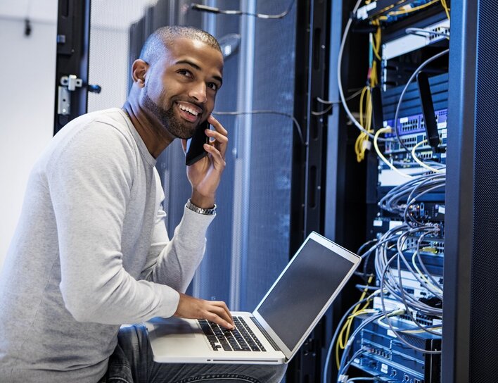 Titelbild Master Cyber-Security Ein Mann mit Laptop spricht lächelnd am Telefon, während er in einem Serverraum an Netzwerktechnik arbeitet.
