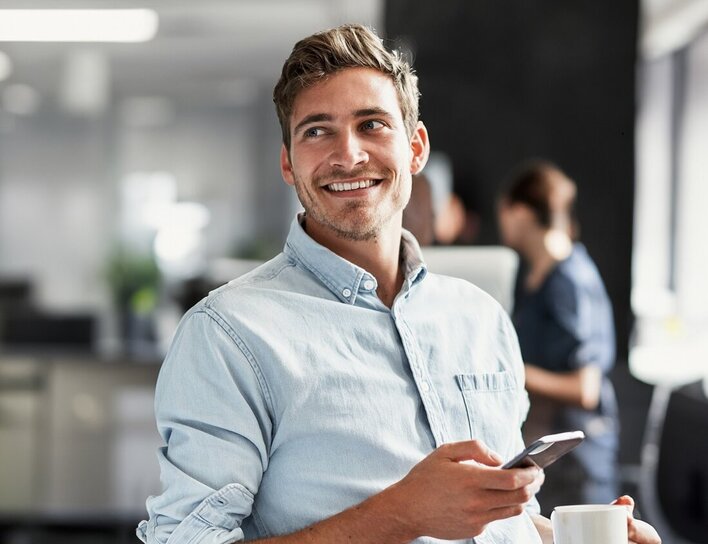 Titelbild Bachelor Betriebswirtschaftslehre Ein junger Mann in heller Kleidung hält Smartphone und Kaffeetasse in einem modernen Büro und blickt lächelnd zur Seite.